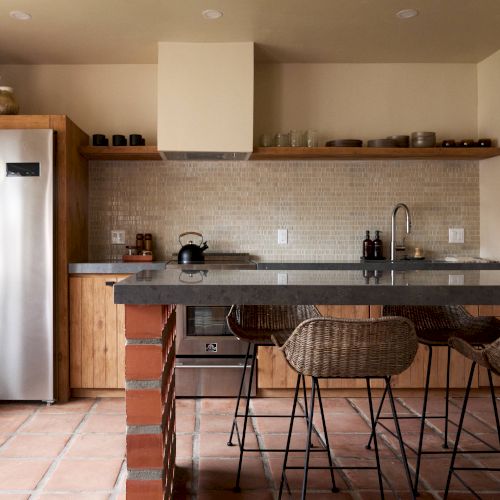 A modern kitchen with a stainless steel refrigerator, tiled backsplash, wooden cabinets, and a counter with wicker chairs.