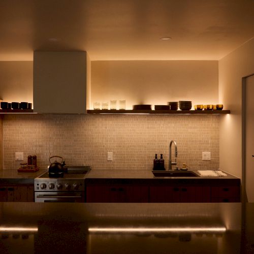 A modern kitchen with under-cabinet lighting, a stove, sink, and open shelves holding dishes and glasses. The decor is minimalist and warm.