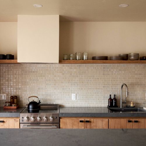 A modern kitchen with a stovetop, oven, black kettle, wooden cabinets, a sink, and a tiled backsplash features open shelving with various items.