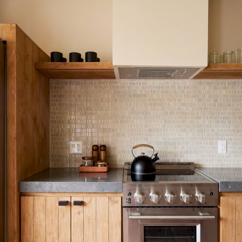 A modern kitchen with wooden cabinets, a stovetop with a kettle, a refrigerator, and shelves holding kitchenware, including glasses and containers.