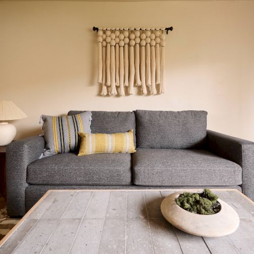 A cozy living room with a gray sofa, colorful pillows, a wooden coffee table, and a macramé wall hanging above. A side table holds a lamp.