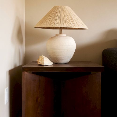 A wooden corner table with a beige lamp and a seashell decoration placed on it, set against a neutral-colored wall.