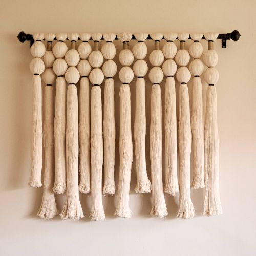 The image features a decorative wall hanging made of suspended, textured beige strands and large beads, creating a visually pleasing pattern.