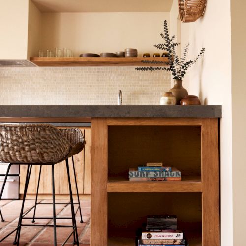 The image shows a cozy kitchen with a wooden island featuring open shelving with books, a bar stool, and some decor items including a vase on the countertop.