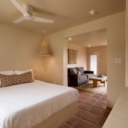 The image shows a cozy bedroom with a bed, a ceiling fan, and an adjoining living area with a sofa and a coffee table in a neutral color palette.