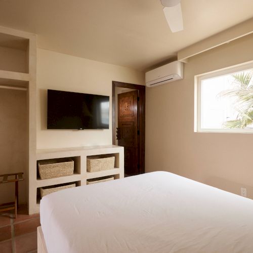 A small, neatly arranged bedroom with a bed, wall-mounted TV, ceiling fan, open closet, and a window showing a partial view of palm trees outside.