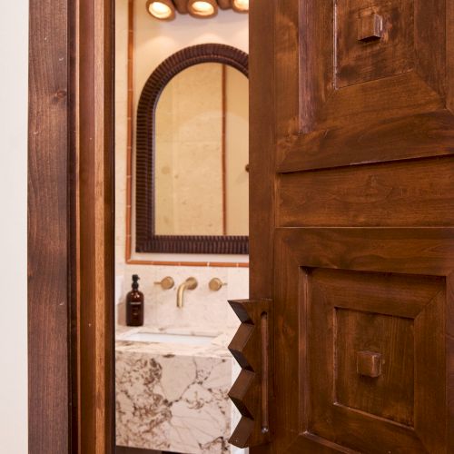 A wooden door opens to reveal a bathroom with a marble sink, a framed mirror, lighting, and various bathroom accessories.