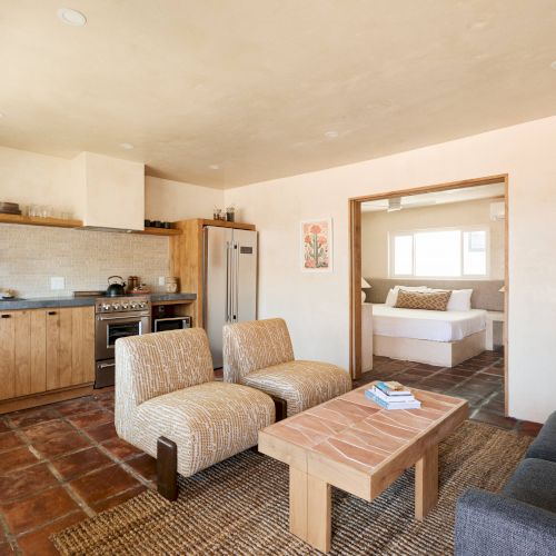 This image shows a cozy, rustic-style living area with a kitchen, two chairs, a sofa, a coffee table, and a view into a bedroom.