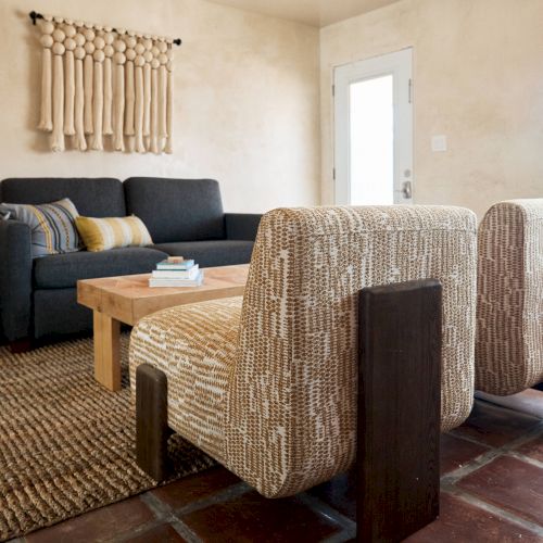 A cozy living room with a dark sofa, patterned chairs, a wooden coffee table, a textured rug, and wall decor, facing a door and window with natural light.