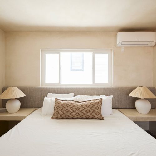 A minimalist bedroom with a large bed, two bedside tables with matching lamps, a decorative pillow, a window, and an air conditioner on the wall.