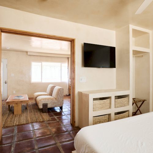 The image shows a cozy living space with tile flooring, wicker storage, a mounted TV, a bed, and a coffee table with two chairs in an adjacent room.