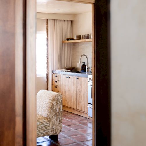 The image shows a view through a door into a room with a kitchen area, wooden cabinets, a sink, and a cozy armchair on tiled flooring.