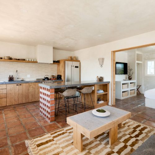 A modern open-plan living space with a kitchen, island, sofa, coffee table, and connected room, featuring tiled floors and bright natural light.