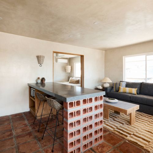 The image shows a cozy open-plan living area with a kitchen and a lounge. It features a sofa, coffee table, kitchen island, and bar stools.