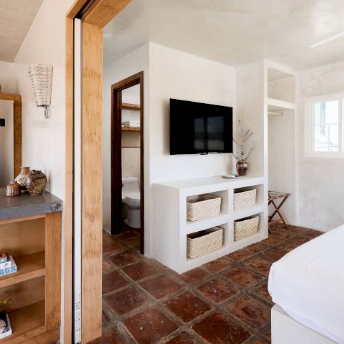 A small, modern room with a kitchen, bed, TV on the wall, shelves with baskets, and a doorway leading to a bathroom.