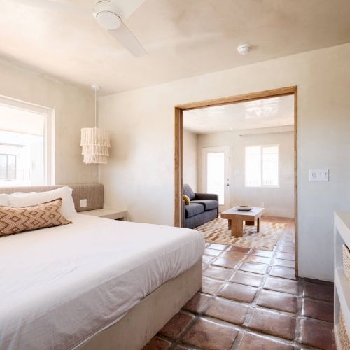 The image shows a bright, minimalist bedroom with a large bed, leading to a living area with a sofa and coffee table, all with tiled flooring.