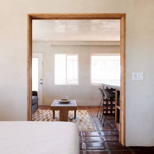 The image shows a bedroom leading to a dining area with a table and chairs, featuring minimalist furnishings and natural light from two windows.