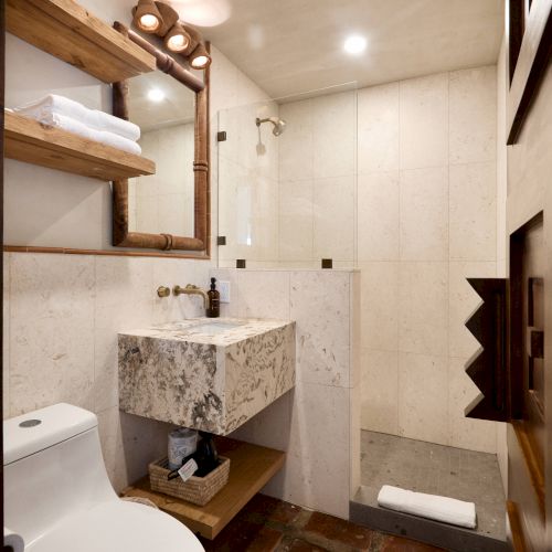 A modern bathroom features a toilet, a floating marble sink with a mirror above it, shelves, and a glass-enclosed shower area.