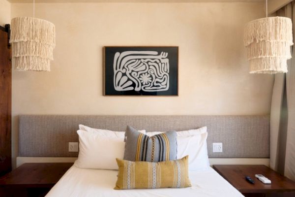 A bed with decorative pillows is between two wooden nightstands, featuring hanging lights and abstract art above the headboard, ending the sentence.