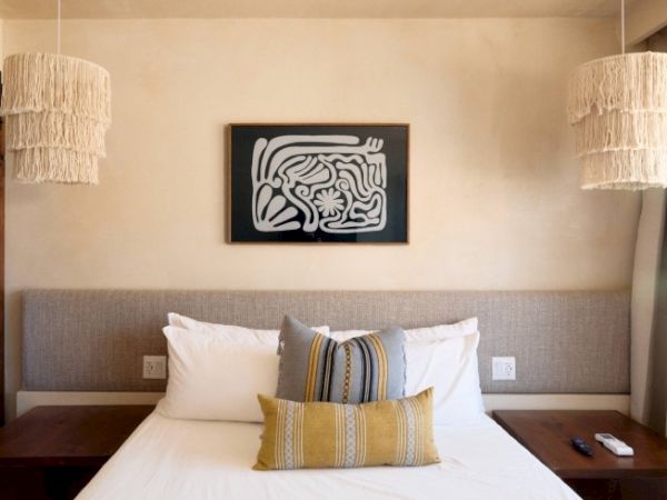 A bed with decorative pillows is between two wooden nightstands, featuring hanging lights and abstract art above the headboard, ending the sentence.