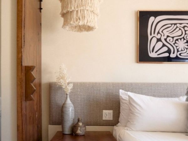 The image shows a stylish bedroom corner with a bed, a wooden nightstand, decorative vases, a woven hanging light, and framed wall art.