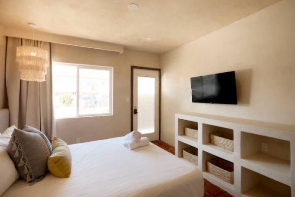 A neatly arranged bedroom with a bed, pillows, a wall-mounted TV, window, door, and built-in shelves with baskets beneath the TV.