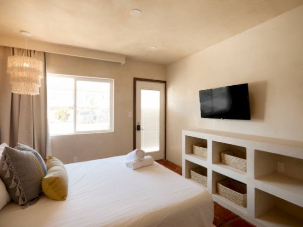 A neatly arranged bedroom with a bed, pillows, a wall-mounted TV, window, door, and built-in shelves with baskets beneath the TV.