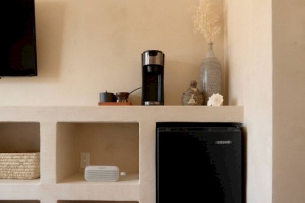 The image shows a shelf with a coffee maker, decorative items, and a small fridge, along with a TV mounted on the wall.