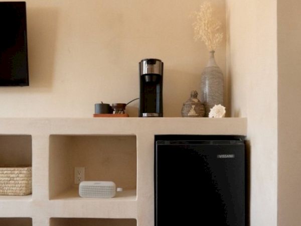 The image shows a shelf with a coffee maker, decorative items, and a small fridge, along with a TV mounted on the wall.