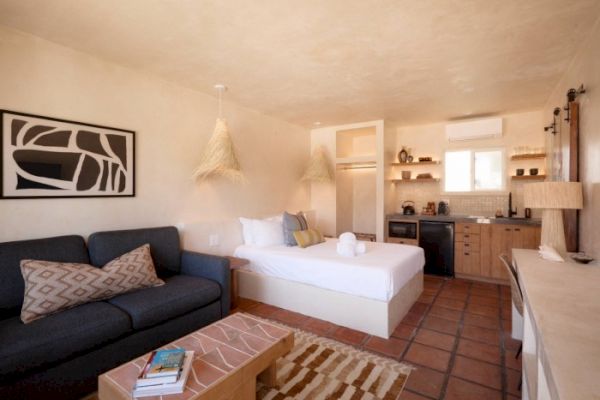 A cozy studio apartment with a bed, couch, coffee table, kitchenette, and rustic decor. Towels are placed on the bed, and shelves hold various items.