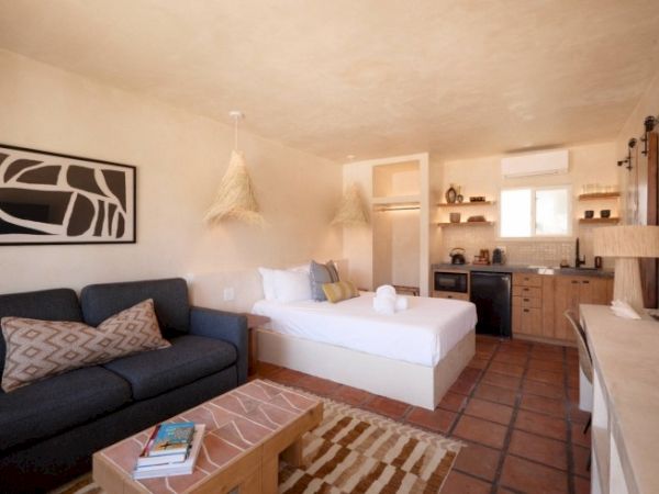 A cozy studio apartment with a bed, couch, coffee table, kitchenette, and rustic decor. Towels are placed on the bed, and shelves hold various items.