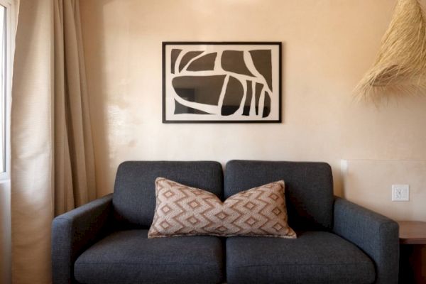 A gray couch with a patterned pillow is in the center, under a black and white abstract wall art. Beige walls and a window with curtains.