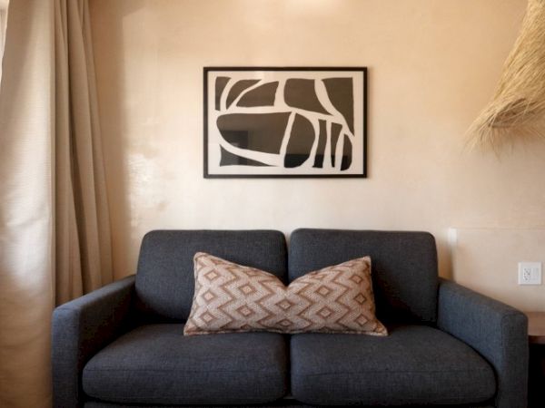 A gray couch with a patterned pillow is in the center, under a black and white abstract wall art. Beige walls and a window with curtains.