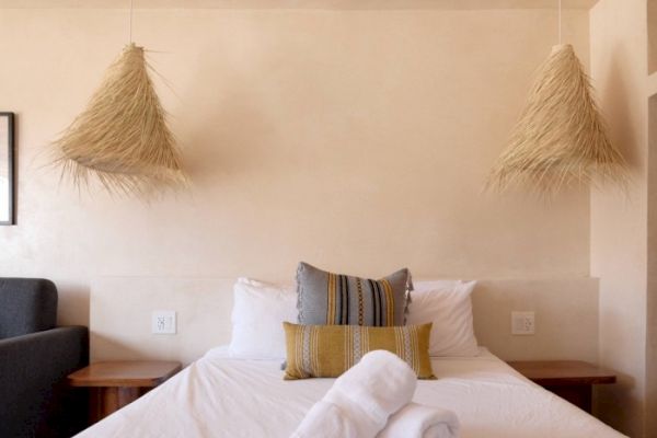 A neatly made bed with white linens, two decorative pillows, rolled towels, and hanging straw lamps on each side in a cozy, minimalist room.