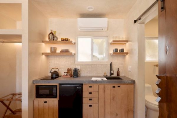 This image shows a small kitchen with wooden cabinets, a countertop, a microwave, a small refrigerator, open shelves, and a window.