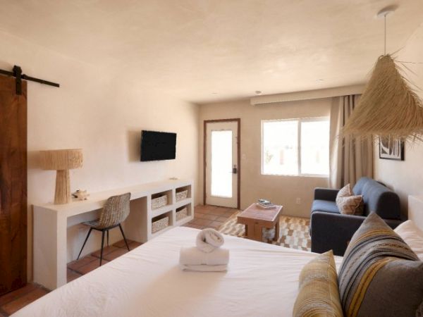 A cozy, minimalistic hotel room with a bed, sofa, TV, desk, and a chair. There’s also a window, a door, and decor elements like lamps and a rug.