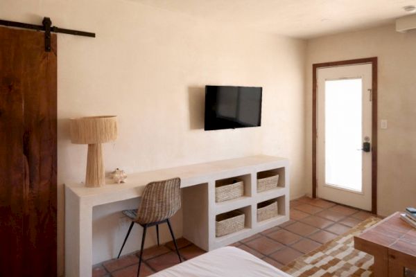 A minimalistic room with a desk, chair, baskets, lamp, wall-mounted TV, a sliding wooden door, tiled floor, and a door with a glass panel to the outside.