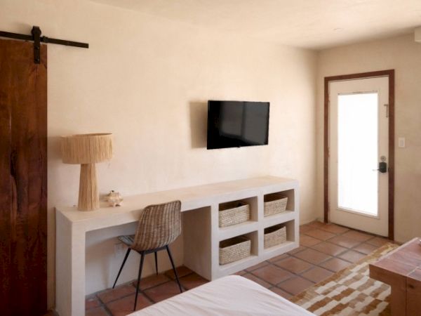 A minimalistic room with a desk, chair, baskets, lamp, wall-mounted TV, a sliding wooden door, tiled floor, and a door with a glass panel to the outside.