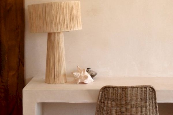 A minimalist desk setup with a tall woven lamp, a seashell, and a small bowl, accompanied by a woven chair.