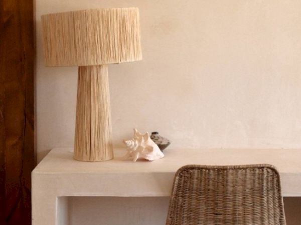 A minimalist desk setup with a tall woven lamp, a seashell, and a small bowl, accompanied by a woven chair.