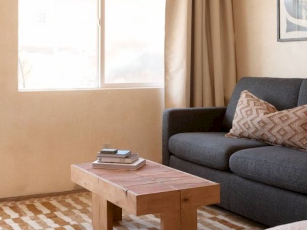 A cozy living room with a dark gray sofa, patterned pillows, a wooden coffee table with books, a rug, a window, and beige curtains.