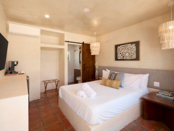 A cozy hotel room with a bed, rolled towels, wall art, a bathroom, a wall-mounted TV, and a small wooden table.