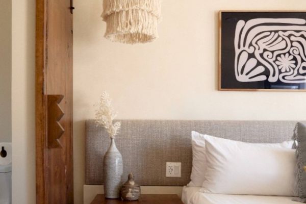 A cozy bedroom with a headboard and a nightstand holding a vase and a small decorative item, a framed abstract artwork, and a hanging woven light.