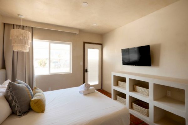 This image shows a bedroom with a bed, pillows, a wall-mounted TV, a window, a door, shelves with baskets, and rolled towels on the bed.