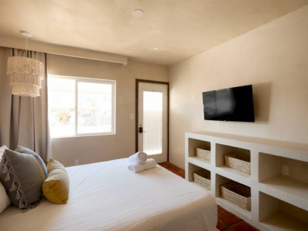 This image shows a bedroom with a bed, pillows, a wall-mounted TV, a window, a door, shelves with baskets, and rolled towels on the bed.
