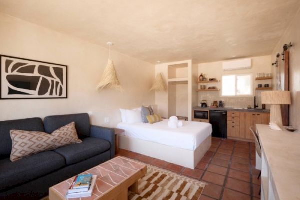 A cozy studio with a bed, sofa, and minimal kitchen. Stylish decor includes a wall art piece, pendant lights, and a rug on tile flooring.