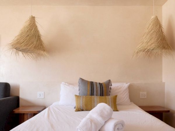 A minimalist bedroom features a neatly made bed with two pillows, rolled towels, and two hanging straw lampshades, flanked by side tables.
