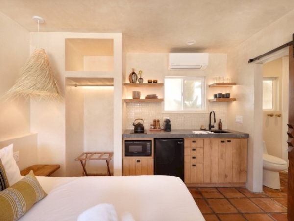 A cozy, rustic-style room with a bed, kitchenette, open shelves, wooden cabinets, a bathroom, and warm-toned decor and lighting.