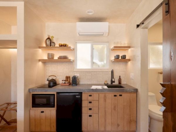 A small, modern kitchen with wooden cabinets, a black mini-fridge, microwave, shelves, and a window. A bathroom is visible through a sliding barn door.
