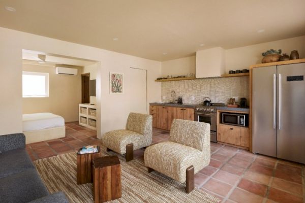 A modern living area with a kitchen, two patterned chairs, a sofa, wooden coffee tables, and a view into a bedroom with a bed and window.